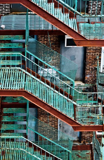 A close-up photo of a weathered teal-and-rust fire escape zigzagging down a brick apartment building, shot at a steep diagonal angle.
