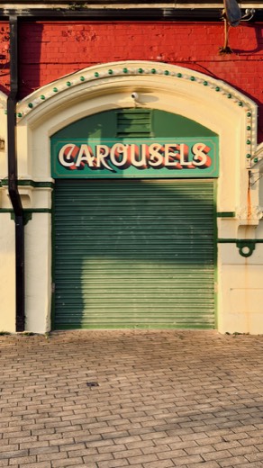 A photo of a shuttered green roll-down door beneath an arched cream facade with a vintage sign reading "Carousels" on a red brick wall.
